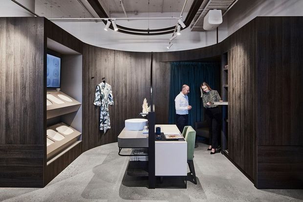 A sophisticated retail showroom or commercial display space with curved dark timber-look wall panelling, integrated illuminated product shelving, a central display island and consultation desk, exposed ceiling services, track lighting, and polished concrete flooring. Two people stand near the rear, with a garment displayed on the wall and a deep blue curtain accent.
