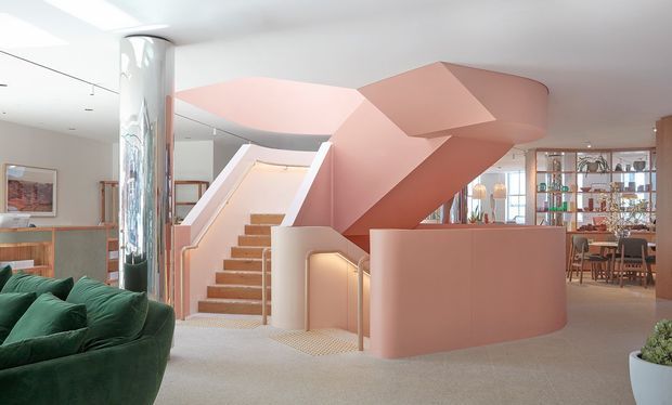 Interior view of a highly sculptural pink staircase and stair enclosure forming the focal point of an open-plan space. The stair has broad timber treads, pale integrated side walls, concealed warm lighting, and a dramatic angular soffit or canopy. A reflective cylindrical column, deep green sofa, display shelving, dining area, artwork, and neutral carpet are also visible. The overall design is contemporary, playful, and gallery-like with strong geometric forms.