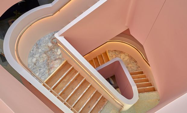 A dramatic overhead view of a sculptural multi-flight stairway with sweeping curved balustrades and pink plaster walls. Warm integrated strip lighting traces the stair edges and curves, while the stairs combine timber-toned treads with pale stone-patterned surfaces and a patterned tiled landing. The composition highlights a sculptural, flowing stairwell design.