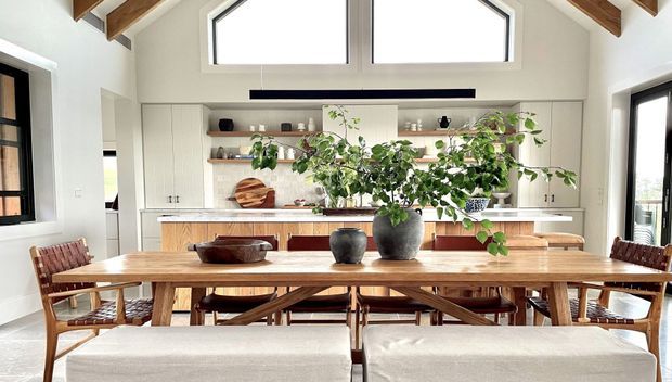 Bright open-plan dining and kitchen interior with a long solid timber dining table, woven timber dining chairs, upholstered bench seating, white built-in cabinetry, open timber shelves, and a light timber kitchen island. Exposed timber ceiling beams and high clerestory windows create an airy, warm space, with greenery and ceramics adding natural accents.