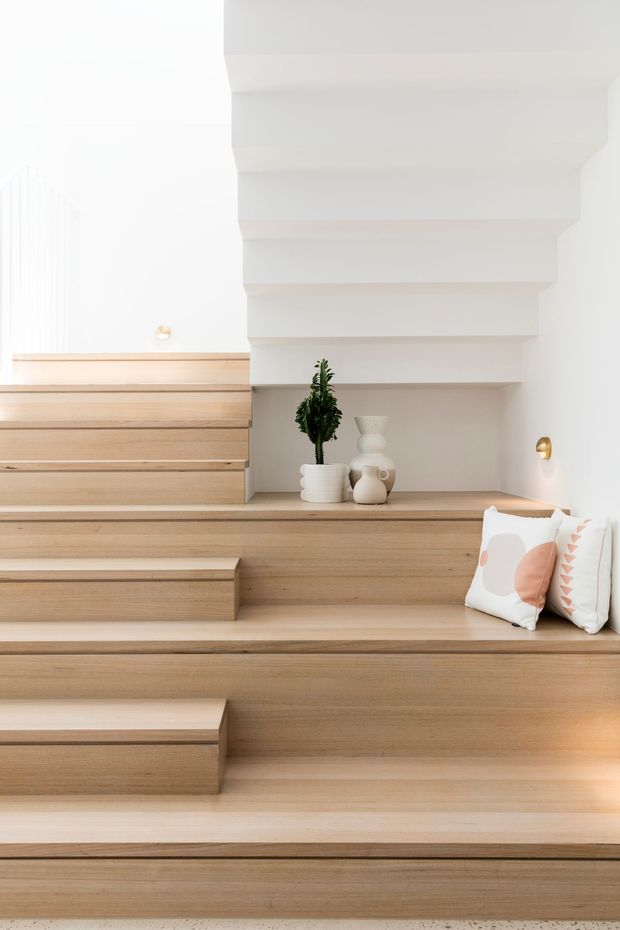 Bright, minimalist interior stairway with broad light timber-clad steps forming tiered built-in seating. White walls and a sculptural stepped ceiling or bulkhead create a layered architectural backdrop, with a recessed display ledge holding plants and neutral ceramic vessels. Soft cushions and discreet wall lighting add warmth and comfort.