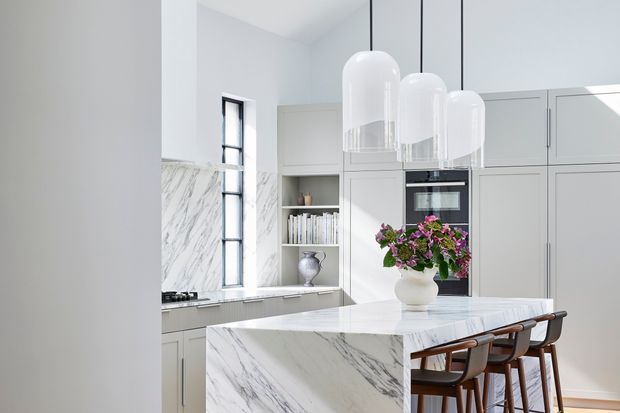 Bright, refined contemporary kitchen featuring a large white marble island with waterfall sides, pale grey built-in cabinetry, a marble backsplash, integrated appliances, open shelving, and three oversized white pendant lights. Timber-framed counter stools and a floral arrangement add warmth to the otherwise light, neutral scheme.
