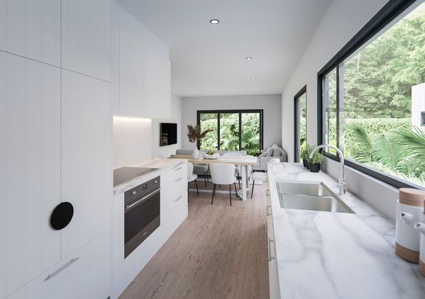 Bright contemporary kitchen with a long white island and integrated cabinetry, marble-look benchtops, stainless steel appliances, a double sink, and large black-framed windows overlooking dense greenery. A dining area is visible beyond the kitchen, with light timber flooring and recessed ceiling lighting.
