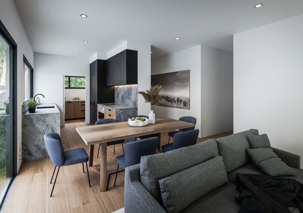 Open-plan contemporary interior combining a dining area, kitchen and living room. A timber dining table with blue upholstered chairs sits in the centre, with a large grey sectional sofa in the foreground. The kitchen includes dark wood cabinetry, a pale stone or marble-look splashback, and a mottled stone island or bench. White walls and recessed ceiling lights create a bright, clean setting, with large windows along the left side.