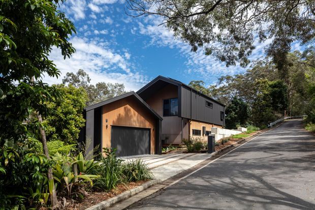 Exterior view of a contemporary multi-level house on a steep, tree-lined suburban site. The building features dark vertical cladding, warm timber garage and gable panels, sharply pitched intersecting roof forms, and a concrete driveway leading to a single garage. Dense established vegetation surrounds the property.
