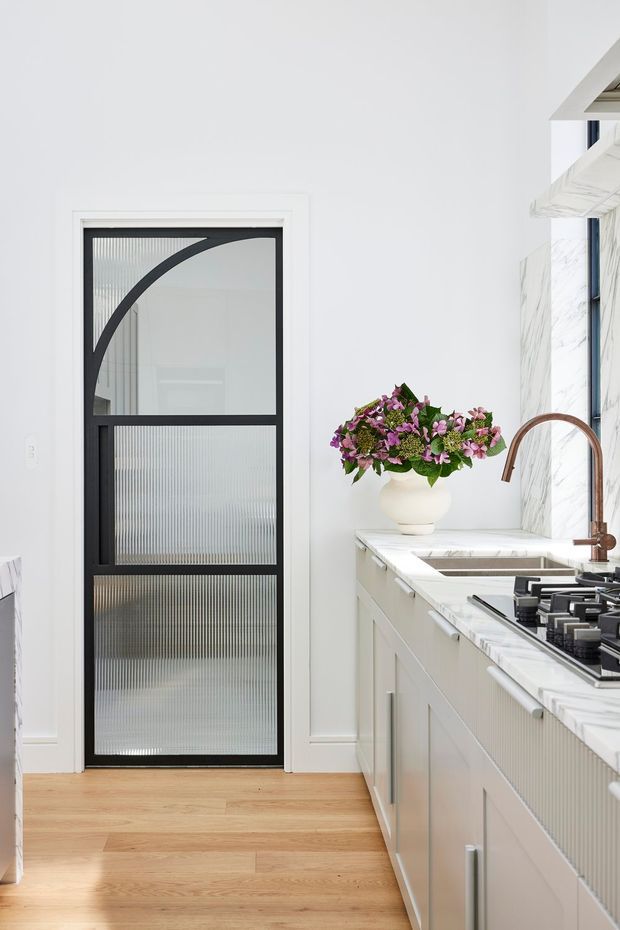 Bright, refined kitchen interior featuring white cabinetry, a veined white stone benchtop and splashback, timber flooring, a gas cooktop, and a curved copper-toned tap. A black-framed reeded-glass door with an arched decorative detail provides a strong architectural feature, while a floral arrangement adds colour.
