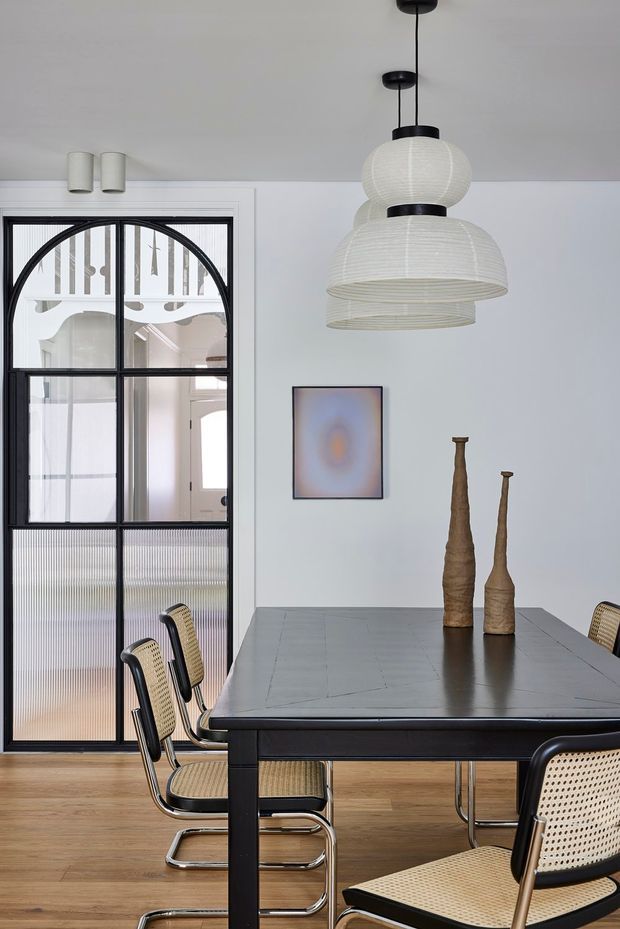 A refined dining room with a large dark rectangular dining table surrounded by woven cane-and-black-frame cantilever chairs. A sculptural cluster of white ribbed pendant lights hangs above the table, while a black-framed glazed internal door with an arched upper detail opens to a bright adjoining space. Pale walls, timber flooring, minimalist artwork, and tall neutral ceramic vessels create a restrained contemporary interior.
