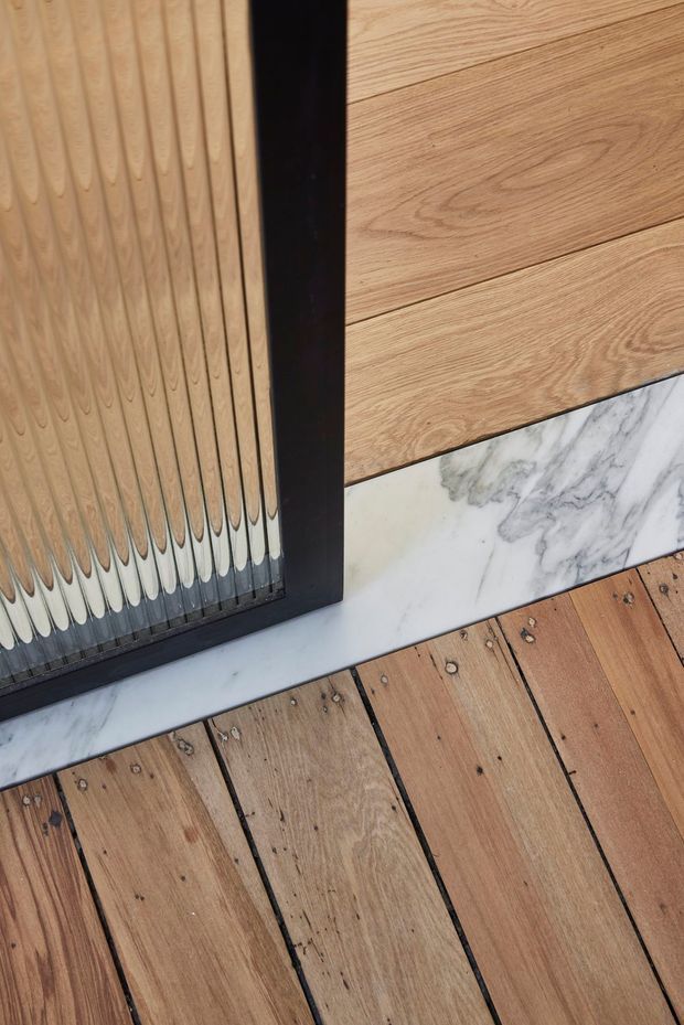 Close-up architectural detail showing warm timber decking with visible fasteners, a black-framed textured glass panel or door, horizontal timber cladding, and a contrasting white-grey veined marble threshold or trim. The image emphasizes material junctions and refined detailing.