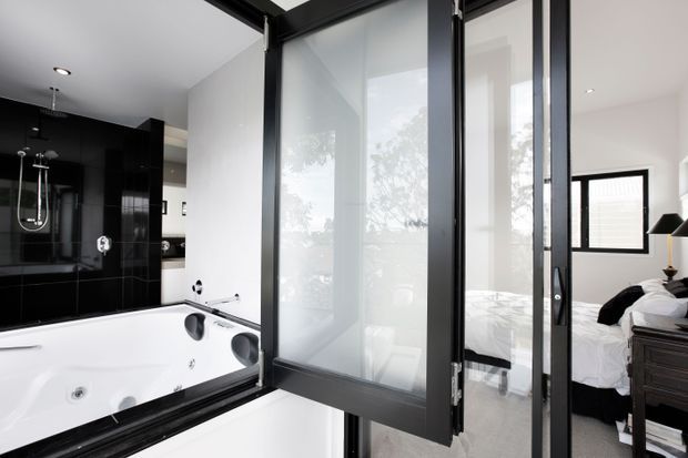 A contemporary bathroom with a large white spa bath and glossy black tiled feature wall, viewed through a black-framed glazed partition and door. A bedroom with a white bed and dark furniture is visible directly beyond the bathroom, indicating an ensuite connection. The space has a bright, high-contrast black-and-white palette and clean-lined fittings.