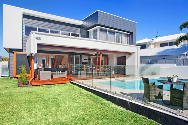 A bright contemporary multi-level residential home viewed from the rear, featuring white and charcoal cladding, extensive glazing, a covered outdoor living area, glass pool fencing, and a rectangular swimming pool beside a lawn and outdoor dining area.