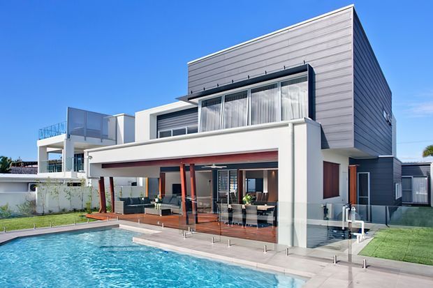 Contemporary multi-level residential home with a strong geometric form, grey horizontal cladding, white rendered walls, extensive glazing, and a covered outdoor entertaining area opening onto a swimming pool. The image is captured from the landscaped pool area and highlights the rear facade and outdoor living connection.