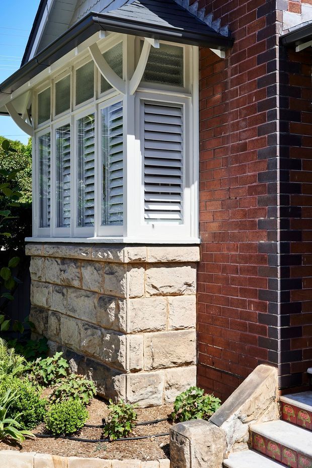 Close-up exterior view of a traditional brick house corner featuring a projecting bay window with white timber frames, textured upper glazing, louvred shutters, decorative curved brackets, and a sandstone block base. A tiled entry stair and landscaped garden bed are visible in the foreground.