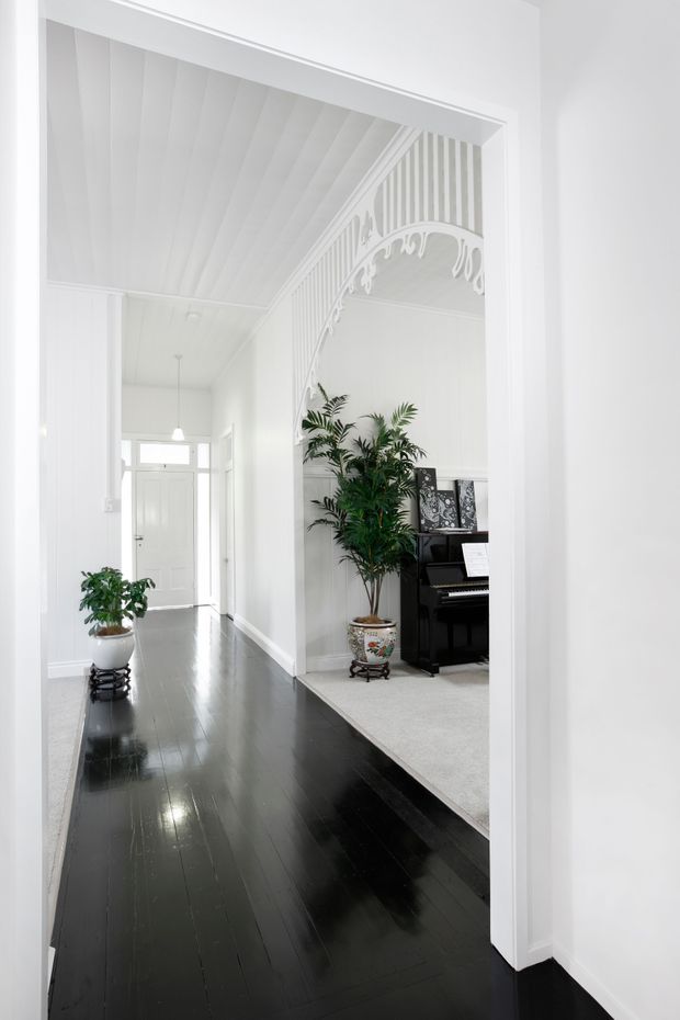 Bright white hallway viewed through a wide doorway, featuring glossy dark timber floorboards, white paneled walls and ceiling, and an ornate decorative arch with fretwork opening into an adjoining room. A black upright piano, framed artwork, and potted plants are visible in the adjacent space. The corridor leads to a white entry door with glazed transom panels.