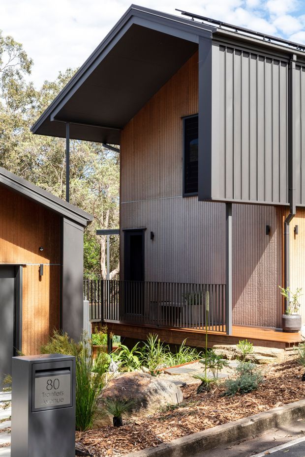 Exterior view of a contemporary multi-level residence set among established trees and landscaped garden beds. The building features a steep asymmetrical gable roof, dark grey vertical metal cladding, warm timber or timber-look vertical wall lining, covered decks and balconies with slim black railings, and strong projecting eaves. A modern dark mailbox and address plaque are visible in the foreground.