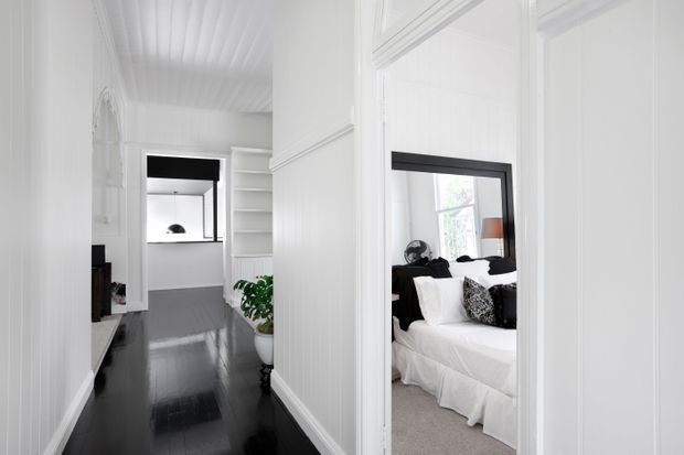 Bright white traditional-style interior showing a hallway with glossy dark flooring, wall panelling, built-in shelving, and a view into a furnished bedroom through a wide doorway. The bedroom contains a white upholstered bed with layered cushions and dark accents. White timber detailing and a fluted ceiling create a refined, high-contrast scheme.