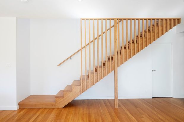 Bright minimalist interior featuring a modern timber staircase with open vertical balustrading, white walls, a white door, and warm timber flooring. The stair rises diagonally across the room and includes a simple timber handrail and central structural post.