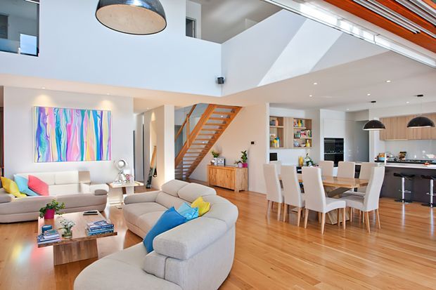 Bright open-plan contemporary home interior with a double-height living area, mezzanine-style upper level, timber staircase with orange-toned handrails, polished timber flooring, a white kitchen, dining area, and large modular sofas. Colourful abstract wall art and cushions add vivid accents to the otherwise neutral scheme.