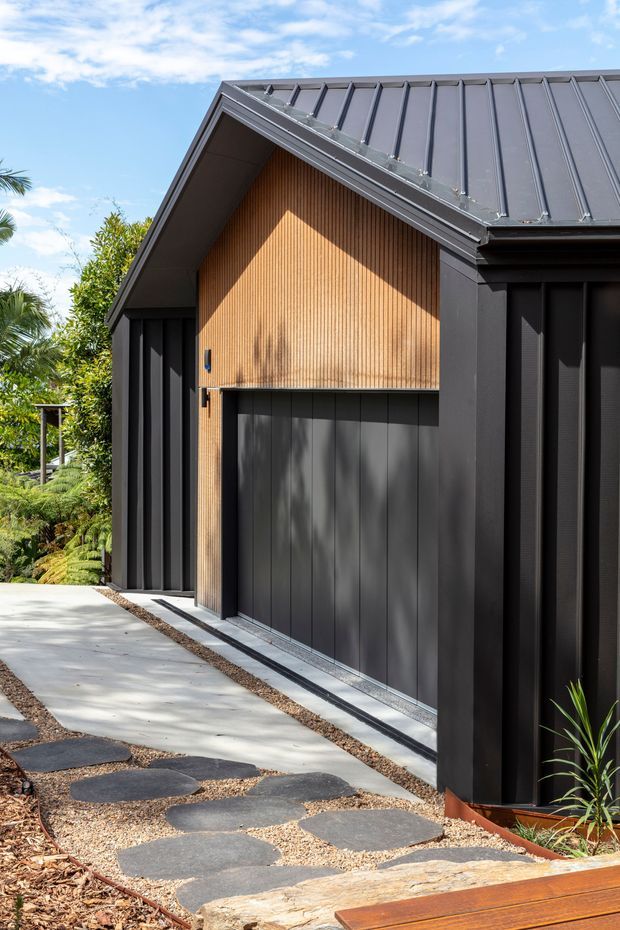 Contemporary dark-clad house exterior with a steep gabled roof, vertical timber feature cladding, and large dark garage doors. A concrete path with irregular stone stepping slabs leads through landscaped greenery toward the entry and garage.