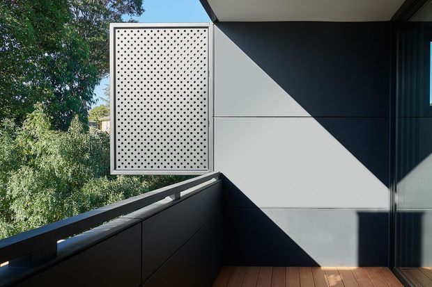 A close-up exterior view of a contemporary building facade from a balcony or terrace. The composition features dark charcoal wall cladding with clean horizontal panel joints, a perforated privacy screen in a light grey frame, dark metal balustrading, and timber decking. Strong sunlight creates crisp triangular shadows across the facade, with leafy vegetation visible beyond the terrace.