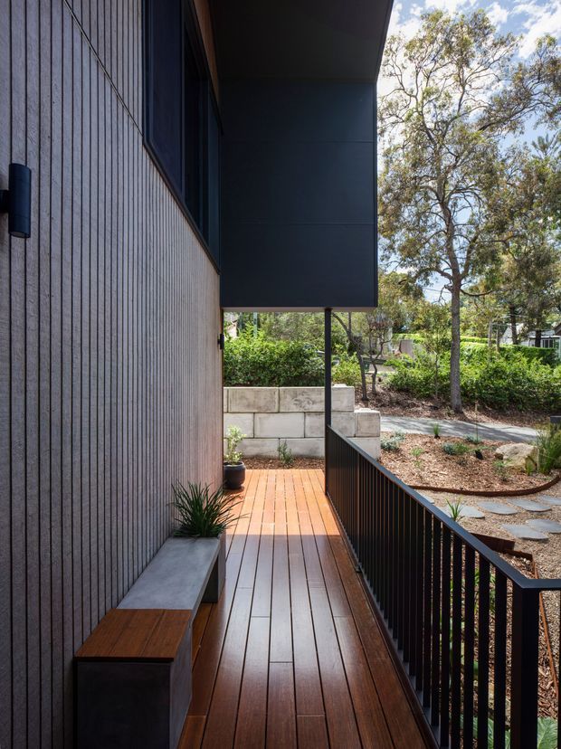A narrow elevated timber deck or external walkway runs alongside a contemporary home. The facade is clad in vertically ribbed grey fibre-cement or composite panels, with dark metal soffits, walls, and balustrading. Built-in concrete and timber bench seating, potted plants, landscaped garden beds, and a concrete block retaining wall are visible. The walkway overlooks a leafy suburban landscape.
