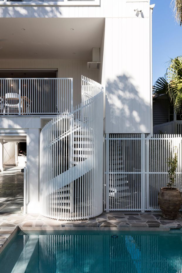 A bright contemporary multi-level residential exterior centered on a sculptural curved white staircase enclosed by vertical white metal balustrades. White vertical cladding, an upper balcony, shaded outdoor areas, patterned stone paving, and a turquoise swimming pool define the scene, with tropical planting at the edges.