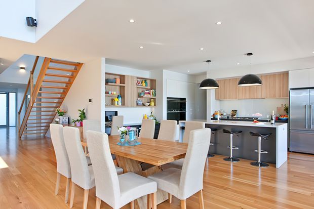 Bright open-plan kitchen and dining area with a long timber dining table, upholstered dining chairs, a contemporary kitchen island with stools, integrated shelving, stainless-steel appliances, and a timber open-riser staircase. Recessed ceiling lighting and black pendant lights add contrast to the light, airy interior.