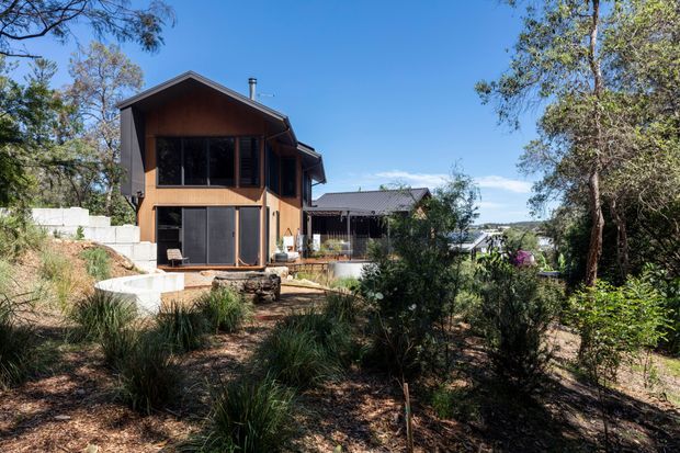 Exterior view of a contemporary multi-level home set within a landscaped, wooded site. The house features warm timber cladding, dark-framed glazing and doors, projecting gabled roof forms, and a partially elevated design responding to the sloping terrain.