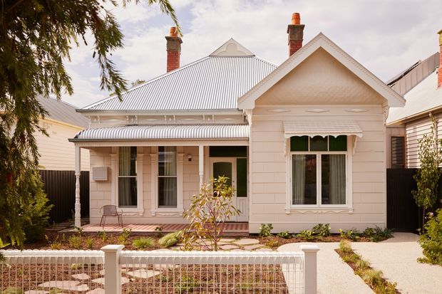 Front exterior view of a renovated single-storey weatherboard house with a gabled roof, corrugated metal roofing, decorative gable detailing, brick chimneys, verandah, timber-framed windows, and a low front fence. The facade is painted in a light cream/off-white palette with a landscaped front garden and paved entry path.