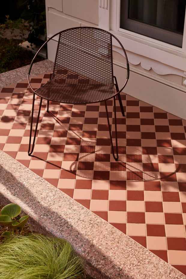 A sunlit outdoor patio beside a white rendered building, featuring a black metal mesh lounge chair on a red-and-cream checkerboard tiled floor. Ornate white trim is visible around the window and wall, with a raised pink-speckled stone garden or terrace edge in the foreground and planting below. Strong shadows create a warm, graphic composition.