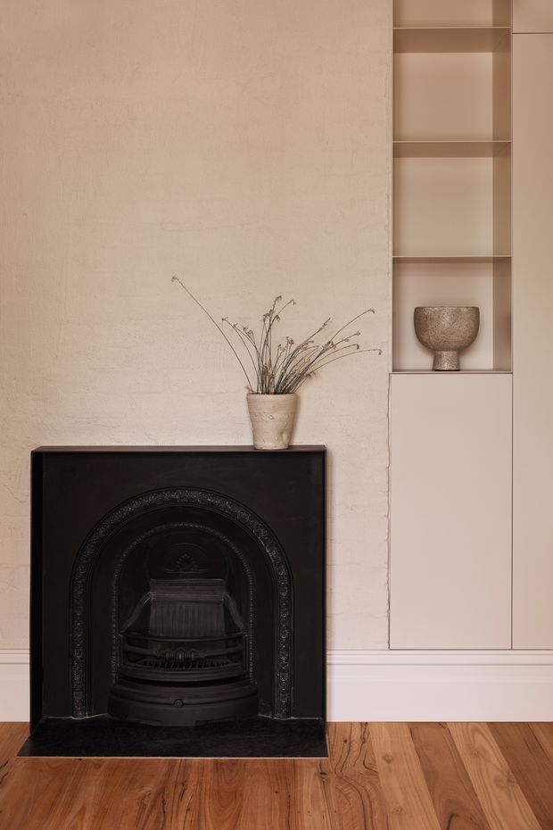 A close-up interior view of a refined fireplace wall. A black cast-iron ornamental fireplace with an arched decorative surround sits against a textured warm off-white plaster wall. To the right is a tall built-in shelving and storage unit in a muted beige finish, displaying a single sculptural vessel. A small ceramic pot with dried branches rests on the mantel, and warm timber flooring grounds the composition. The overall styling is understated, monochromatic and heritage-inspired.