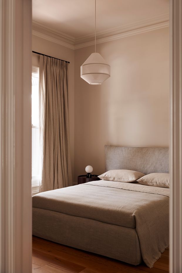 A warm, softly lit bedroom viewed through a doorway. The room features a neatly made upholstered bed with a textured beige headboard and neutral bedding, full-length taupe curtains beside a window, a small dark bedside table with a spherical lamp, a contemporary pendant light, timber flooring, and ornate ceiling cornices and door trim. The overall scheme is calm, refined, and tonal.