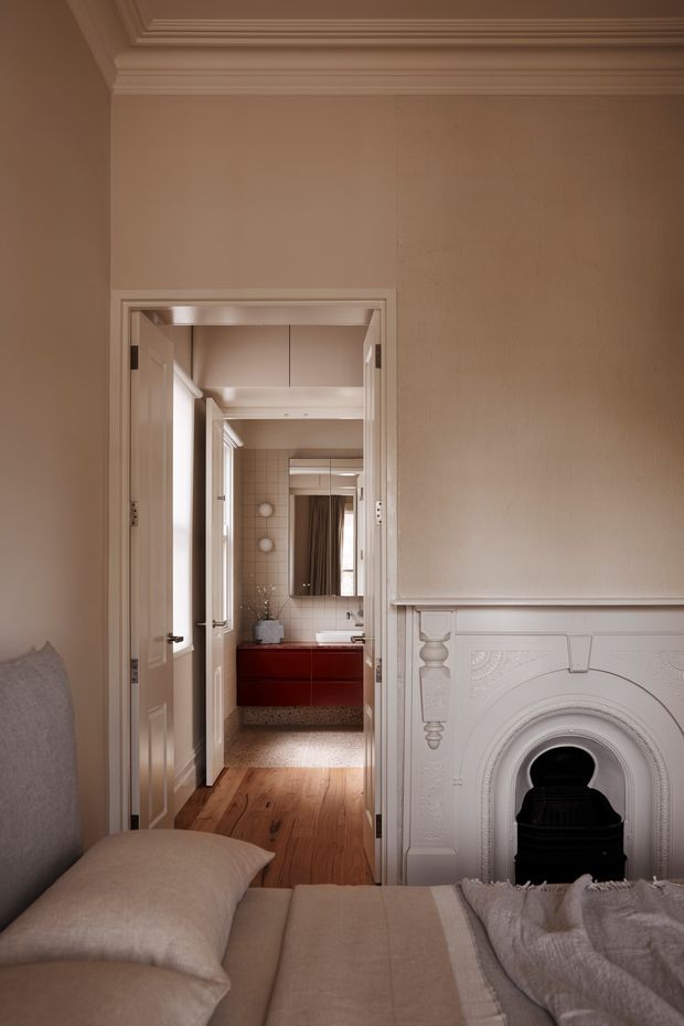 A warmly lit traditional bedroom viewed toward an open doorway leading to a bathroom. The bedroom features a beige upholstered bed with layered neutral bedding, timber flooring, ornate white wall panelling, decorative cornices, and a detailed white fireplace surround with a dark insert. Through the doorway, the bathroom has a red floating vanity, tiled walls, a mirror, and a timber floor transition.