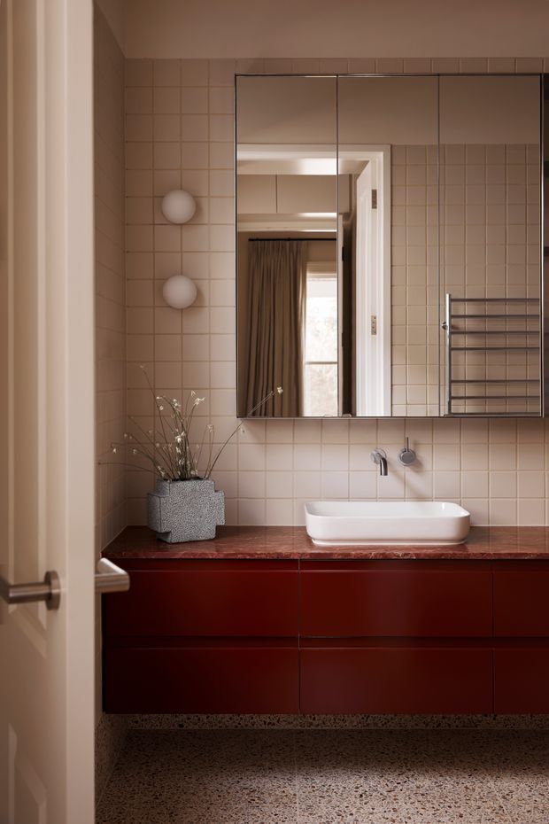 A warm, refined bathroom viewed through a partially open door. The room features a floating deep red vanity with a polished reddish stone benchtop, white vessel basin, wall-mounted tapware, a large mirrored cabinet, pale square tiled walls, a chrome heated towel rail, globe wall lights, and a terrazzo floor. The mirror reflects an adjacent curtained space.