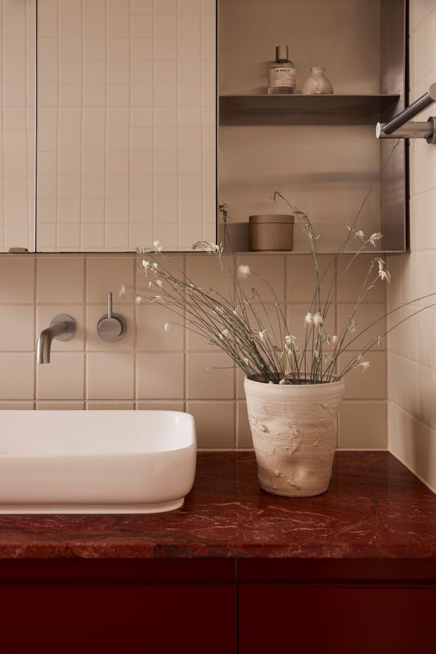 Close-up architectural photograph of a warm, compact bathroom vanity. Cream square wall tiles surround a white inset basin with brushed metal wall-mounted tapware. A dark red-brown marble or natural stone benchtop sits over deep burgundy cabinetry, with a recessed open shelf in a muted taupe finish. A textured ceramic vase with dried grasses adds organic styling.