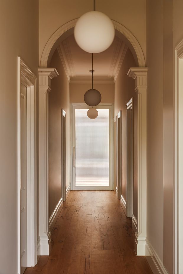 A warm, symmetrical residential hallway with rich timber flooring, softly painted taupe walls, ornate white classical architraves, columns and cornices, and multiple spherical pendant lights suspended along the corridor. Frosted glazing at the far end provides diffused daylight. The space has a refined heritage-inspired character with strong axial perspective.