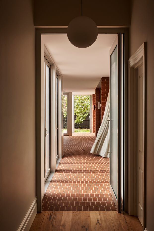 A warmly lit interior hallway leading through glazed doors toward a bright courtyard or garden. The corridor has red brick tiled flooring, timber flooring in the foreground, pale painted walls and trim, multiple glazed doors, a brick feature wall, and a large spherical pendant light. A draped curtain and outdoor greenery are visible at the far end.