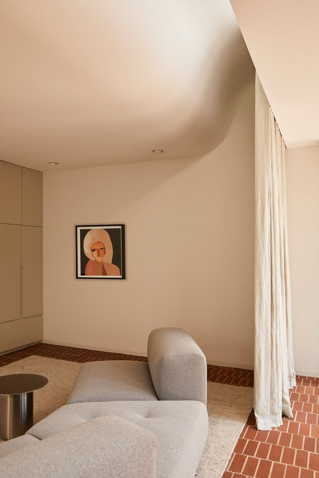 Warm, refined living room interior with a sculptural cream upholstered sectional sofa, round metal side table, pale textured carpet, terracotta brick flooring, full-height sheer curtains, built-in beige cabinetry, and a curved ceiling transition. A framed artwork provides a small accent on the otherwise softly neutral wall.