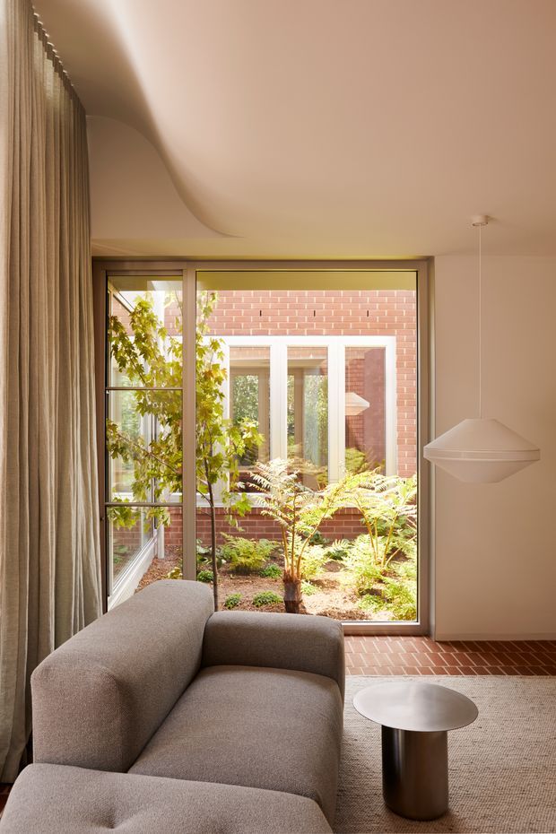 A warm, refined living room with a softly sculpted ceiling, full-height curtains, large glazed doors opening toward a planted courtyard, and a neutral upholstered sectional sofa. A small round side table and suspended pendant light complement the calm interior. Brick paving and an adjoining brick courtyard are visible beyond the glazing.