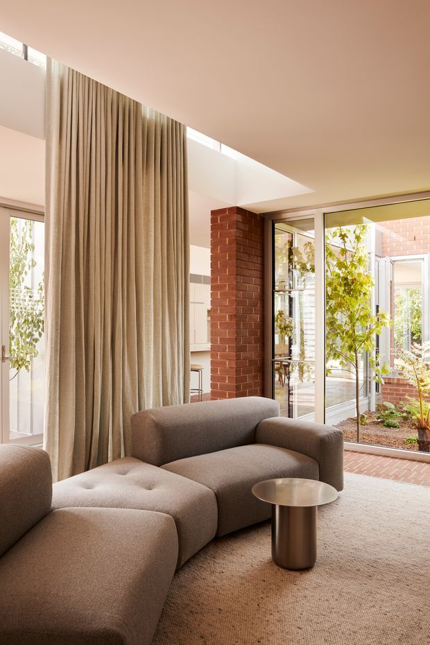 A warm, contemporary living room with a large taupe modular upholstered sofa and chaise, a small round metallic side table, and a neutral textured carpet. Full-height curtains divide the room and frame a bright courtyard-facing glazed wall and doors. A red brick internal column, skylight or clerestory glazing, and views into a planted brick courtyard add architectural interest.