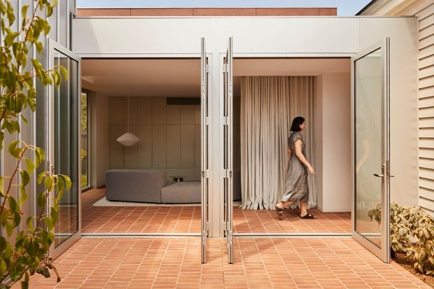 A contemporary indoor-outdoor living area viewed from a terracotta-tiled courtyard or patio. Fully opened glazed bi-fold doors connect the exterior terrace to a lounge with a low grey sofa, tiled feature wall, pendant light, and floor-to-ceiling curtains. Pale rendered walls, timber roofline detailing, and horizontal louvre cladding frame the opening, with planting at the edges.
