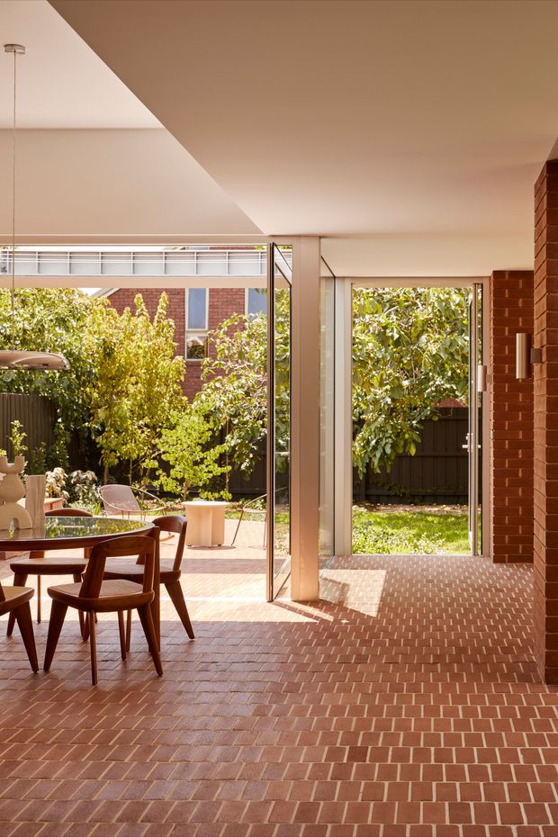 A sunlit indoor-outdoor dining and living area opening onto a leafy courtyard through large glazed doors. The space features extensive red brick flooring continuing toward the garden, exposed red brick walls, warm neutral plaster ceilings, slim framed glazing, and a mid-century-inspired timber dining table and chairs. The courtyard includes paving, planting, and a small outdoor seating area visible beyond the open doors.