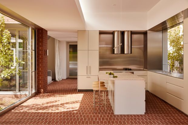 A warm, contemporary kitchen viewed from an open-plan interior, featuring a white island with timber stools, sleek white cabinetry, a dark timber-toned backsplash, integrated appliances, and a large window. Red brick flooring continues through the kitchen and adjacent living area, while exposed red brickwork and extensive glazing add texture and natural light.