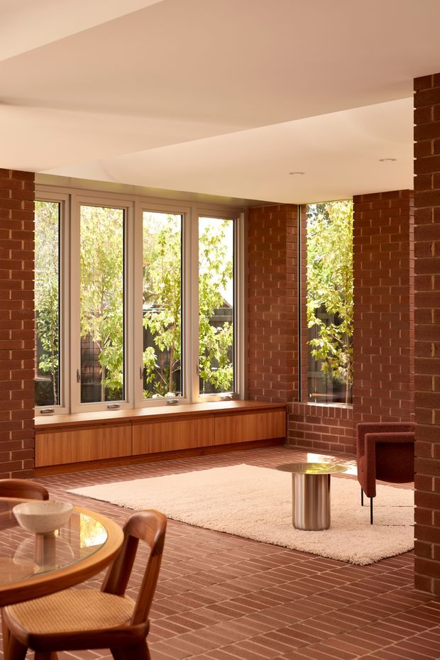 A warm, light-filled open-plan interior with exposed red brick walls and brick flooring. Tall timber-framed windows overlook leafy greenery, with built-in timber window seating and cabinetry beneath them. The space includes a dining table in the foreground and a small lounge area with a burgundy armchair and round metallic side table. A sculptural stepped ceiling and recessed downlights add architectural interest.