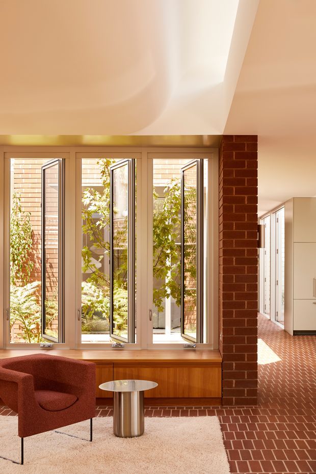 Warm, sunlit contemporary interior centered on a full-height glazed window wall opening toward a leafy courtyard. A built-in timber window seat and cabinetry sit below the windows, beside an exposed red-brick structural column. The room includes a rust-red upholstered lounge chair, small round metallic side table, and pale textured rug. Red brick flooring continues into a bright kitchen or circulation area with white built-in cabinetry. The ceiling has a sculptural curved plaster form and a concealed skylight or clerestory-like opening.