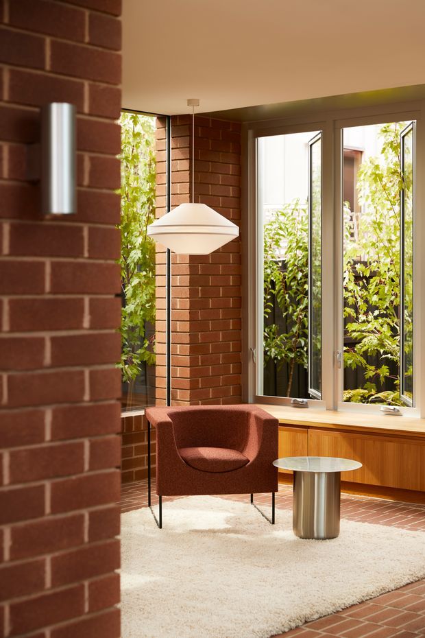 A warm, sunlit modern interior corner with exposed red brick walls, full-height opening windows, and a built-in timber window seat or cabinet. A rust-coloured lounge chair sits beside a small round metal side table on a light textured rug, with a sculptural white pendant and floor lamp providing lighting. Greenery is visible outside through the open windows.