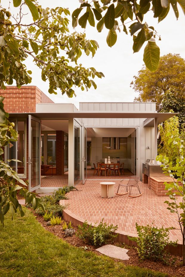 A warm architectural exterior view of a contemporary brick house opening onto a spacious raised brick terrace and landscaped garden. Full-height glazed doors are folded open to connect the terrace with an interior dining and kitchen area. The home combines red brick walls, pale metal-framed glazing, a flat roof, and a covered outdoor entertaining zone, with mature leafy trees framing the composition. Outdoor furniture and a built-in barbecue are visible on the terrace.