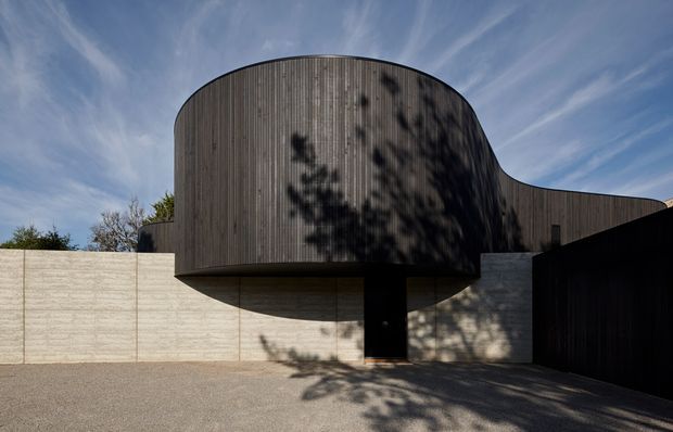 Exterior architectural photograph of a sculptural contemporary building with a rounded, projecting upper volume clad in narrow vertical dark timber boards. The curved form is supported above a pale textured concrete wall, with a dark recessed entry or opening beneath and a large dark garage-like opening to the right. Strong tree shadows fall across the facade and paved forecourt, while a blue sky is visible above.