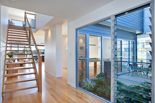 Bright contemporary interior showing an open timber staircase, pale timber flooring, white walls and a large glazed opening with louvred screening. Through the glazing is an enclosed courtyard and a glimpse of a billiards or games area, with abundant natural light and indoor planting.