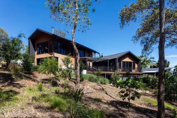 Exterior view of a contemporary timber-clad home set into a sloping, tree-filled site. The house has multiple connected pitched-gable roof forms, dark-framed glazing, covered decks and verandas, and warm brown exterior walls.