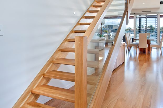 Bright contemporary open-plan interior featuring a timber staircase with open risers, glass balustrade, and substantial timber handrails. The stairway opens toward a dining area with timber flooring, dining furniture, large windows, and horizontal blinds.